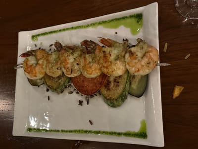 On a dark wooden table, a rectangular white plate holds a shrimp skewer. The skewer includes grilled shrimp, zucchini slices, and a tomato slice, seasoned with herbs. The plate is decorated with lines and dots of green sauce. To the right of the plate is a partially visible wine glass. The location appears to be a restaurant setting, potentially in Tamarindo, Costa Rica.