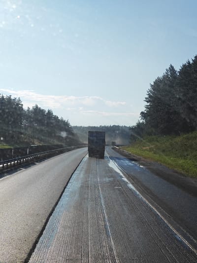 The image captures a highway scene on a sunny day, with a clear blue sky dotted with light clouds. The road appears to be undergoing resurfacing. The foreground shows a freshly paved lane with a textured surface, likely due to recent work. Further down the road, a truck is visible, emitting a faint trail of smoke or dust. On the right side of the road, a dense line of coniferous trees forms a natural barrier, suggesting a rural or semi-rural location. A guardrail runs along the left side of the highway, separating the lanes and the embankment beyond. The overall impression is one of ongoing infrastructure work on a highway surrounded by nature. This scene is located on the highway near Kurumoch, Russia.
