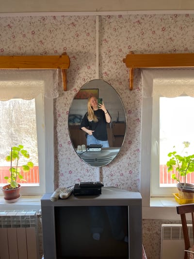 A woman stands in an indoor location, reflected in an oval mirror. She holds a phone, presumably taking the picture. The mirror is mounted on a wall covered with patterned wallpaper. Beside the mirror is a television and several remote controls. Natural light enters through a window near a potted plant. The setting has floral wallpaper. Based on visual analysis and the presence of Russian text, the image may be taken at Чернышово, Russia.