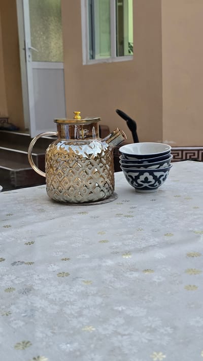The image shows a decorative glass teapot with a gold lid and handle, placed on a tablecloth with a floral pattern. To the right of the teapot, there are three small ceramic bowls stacked on top of each other, each with a blue and white pattern. The scene appears to be set outdoors, possibly on a patio or balcony, with a building and a window visible in the background. The overall impression is one of a relaxed, domestic setting, perhaps prepared for tea time. The image does not provide any specific clues to determine its exact location in Samarkand, Uzbekistan, as such decorative teaware and outdoor seating arrangements are common in many regions.