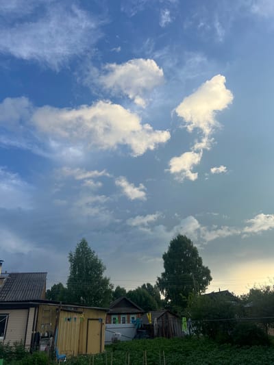 This is a vertical, slightly angled, outdoor photograph taken during the day in what appears to be a rural or semi-rural setting in Pigozino, Russia. The scene is dominated by a bright blue sky filled with scattered white clouds of various shapes and sizes. The sun is not directly visible, but its light illuminates the clouds, giving them a soft glow, and casts a warm light on the horizon.

In the lower portion of the frame, several wooden structures are visible, suggesting a residential area. These structures appear aged, with one building painted yellow and another with a grey, corrugated roof. There are clotheslines strung between poles, with several clothespins attached, indicating recent or ongoing laundry drying. Tall trees with dense green foliage are interspersed with the buildings, adding to the natural feel of the environment. A power line stretches across the scene, supported by poles. The overall atmosphere is peaceful and tranquil, evoking a sense of rural simplicity. The time of day appears to be late afternoon or early evening due to the warm lighting and the angle of the sun. There is no visible text in the image.