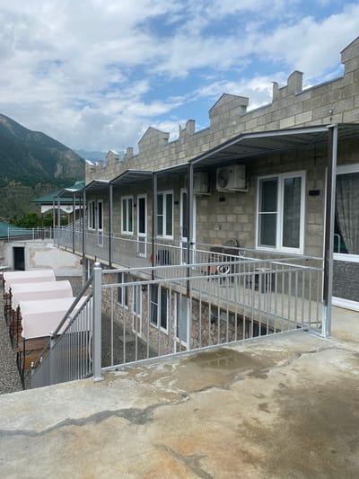 This is a medium-shot, eye-level, outdoor photograph taken in Chalda, Russia. The image features a multi-story building constructed with light-colored cinder blocks and stone. The building has a row of windows with white frames and a metal awning extending from the upper floor. The rooftop is adorned with decorative crenellations, resembling a castle's battlements. In the foreground, a concrete patio with cracks and water stains leads to a metal railing that encloses a walkway. Several white covered lounge chairs are visible on a lower level. In the background, a lush green mountain slopes upwards under a cloudy sky. The overall impression is one of a rustic or resort-like establishment nestled in a mountainous region. There are no people visible in the image.