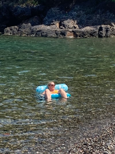 A woman is floating on an inflatable raft in the clear, shallow waters of Pozzarello, Italy. The raft is blue and white with a floral pattern. The woman is wearing sunglasses and an orange swimsuit. She is lying on her back, with her legs extended. The water is calm and transparent, revealing the rocky seabed. In the background, there are rocky cliffs and lush green vegetation. The sun is shining, casting a warm glow on the scene.