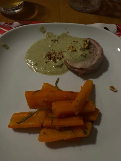 An overhead shot displays a plated meal served on a white, oval-shaped ceramic dish. On the left side of the plate, a cylindrical portion of cooked meat, possibly wrapped in a thin slice of ham or bacon, is presented. It is generously covered in a light green, creamy sauce, which also pools around the base of the meat. Sprinkled over the sauce and the meat are small, chopped pieces of what appear to be nuts, likely walnuts.
To the right side of the plate, a mound of orange, cut vegetables is arranged. These vegetables resemble carrots or sweet potatoes, cut into long, thick sticks or fries, and are garnished with slender green herbs, possibly rosemary or thyme. The lighting suggests an indoor setting, casting distinct shadows from the food items onto the plate, indicating a light source from above and slightly to the left.
The plate rests on a surface, part of which is visible on the left and bottom-left, showing a warm, wooden texture, likely a table. Underneath the left side of the plate, a section of a red and white patterned fabric, possibly a napkin or tablecloth, is visible. Partial views of two drink glasses are seen on the far left of the frame; one in the top-left corner and another in the bottom-left, both containing a yellowish liquid. No text is visible in the image.