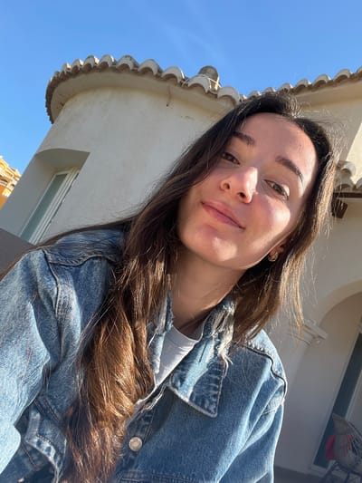 A selfie of a young woman with long brown hair, taken outdoors on a sunny day in El Poble Nou de Benitatxell/Benitachell, Spain. She is wearing a denim jacket over a light-colored shirt and is looking directly at the camera with a slight smile. The background includes a building with a white exterior and ornate roof details, along with a clear blue sky. Part of another building is visible in the lower left corner.