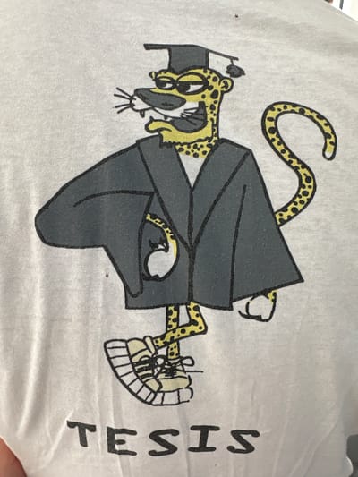 This image is a close-up of a white t-shirt with a printed graphic. The graphic depicts a cartoon leopard wearing a graduation cap and gown. The leopard has yellow fur with black spots, a yellow mane, and black facial features. It is standing on one leg, with its other leg bent, and its tail is curled. The leopard is also wearing sneakers. Below the leopard graphic, the word "TESIS" is printed in black capital letters. The image does not contain any information about the city of Moortown, Ireland.