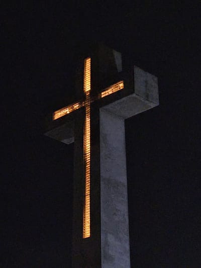 The image shows a concrete cross structure at night, illuminated from within. The cross is a prominent landmark, located in the city of Chillán, Chile, specifically at the entrance of the Universidad de Concepción campus. The structure is dark gray and appears to be made of concrete. Light, with a warm, orange-yellow hue, emanates from rectangular openings that form the shape of the cross. The background is completely black, indicating the nighttime setting and suggesting that the surrounding environment is not visible or is also dark. There are no people or other objects visible in the frame. The focus is solely on the illuminated cross.