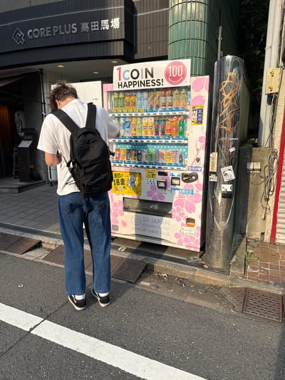 A person with a black backpack stands in front of a pink and white vending machine in Toshima, Japan. The vending machine is stocked with various bottled drinks and features a large graphic of cherry blossoms. Above the vending machine, a sign reads "CORE PLUS 高田馬場," indicating the location is Core Plus Takadanobaba. The person is wearing a white t-shirt, blue jeans, and black sneakers with white socks. They are looking down at the vending machine, appearing to be making a selection. The background shows a building with a dark facade and green tiled architectural elements. The street is paved with asphalt, and there are metal grates on the sidewalk. The lighting suggests it is daytime, with sunlight casting shadows on the ground.