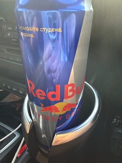 A Red Bull energy drink can is held in a car's cup holder. The can is blue and silver with the Red Bull logo. Text in Cyrillic script is visible on the can. The car's interior is visible in the background, including buttons and a screen. The scene appears to be in Polsko Padarevo, Bulgaria.