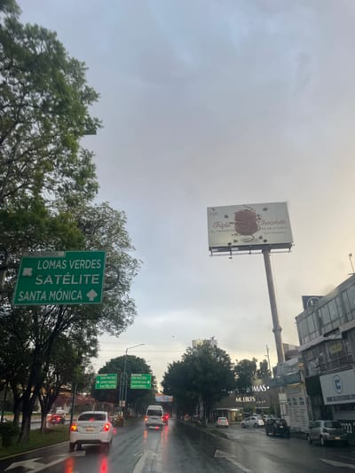 The image captures a view from a vehicle on a wet, overcast day in Tlalnepantla, Mexico, likely on a major thoroughfare. Several green directional signs are visible. The most prominent sign, on the left, indicates "LOMAS VERDES SATÉLITE SANTA MÓNICA". Further down the road, another sign points to "SAN MATEO LOMAS VERDES SATÉLITE" and a separate one indicates "RAQUEL AGUILERA SERDÁN ATIZAPÁN CENTRO SANTA MÓNICA".
A large billboard featuring a stack of chocolate confections and the text "Triple Chocolate EL FAVORITO DE LA CIUDAD VEN A PROBARLO" is prominently displayed on the right side of the road. Adjacent to the billboard, a modern building with multiple stories and windows is visible. Further down the street, a commercial building with the name "THOMAS MUEBLES" is present.
The road is wet and reflective, indicating recent rain, with vehicles driving on it. Several cars are present, including a white sedan and a white van. Trees line the sides of the road, and the sky is a dull, cloudy gray. The overall scene suggests a busy urban environment with clear directional signage for navigation.