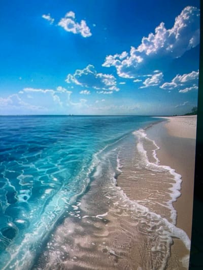 This is a high-angle, full-frame, eye-level shot of a tranquil beach scene under a bright blue sky dotted with fluffy white clouds. The water is a vibrant turquoise, clear enough to see the sandy bottom with ripples of light reflecting on its surface. Gentle waves are washing ashore onto a smooth, light-colored sandy beach. The horizon line is sharp, separating the sea from the sky, with a faint, distant landmass visible on the far right. There are no people, animals, or man-made objects immediately apparent in the scene. The image depicts a serene, natural coastal environment. The location is not Navahrudak, Belarus.