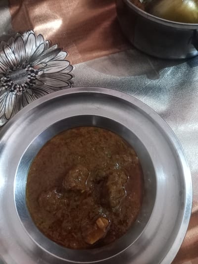 The image shows a close-up, overhead shot of food in metal dishes, likely taken in Orai, India. A shallow, round metal plate holds a brown, gravy-based dish containing several chunks of what appears to be meat. The consistency of the gravy is thick. A second, similar metal container sits partially visible in the upper left corner, also appearing to contain a gravy-like food item, with a yellow hue. The dishes are placed on a patterned surface, possibly a tablecloth or mat. The pattern features floral designs.