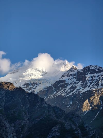 This is a vertical, medium-range, eye-level shot of the majestic mountains in the city of Gemur, India, under a clear blue sky. The image features a dramatic contrast between the dark, jagged peaks in the foreground and the snow-capped mountains in the background. The foreground consists of rugged, rocky slopes, partially covered in dark vegetation, casting deep shadows. The mid-ground is dominated by the imposing, snow-covered mountains, some parts of which are illuminated by the sun, highlighting their icy surfaces and deep crevasses. Wispy clouds partially obscure the highest peaks, adding a sense of depth and mystery to the scene. The sunlight catches the snow, creating bright highlights and casting subtle shadows across the glacial formations. The scene conveys a sense of scale and natural grandeur, characteristic of high-altitude mountain landscapes.