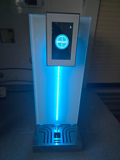 The image shows a close-up, eye-level, medium shot of a modern, illuminated dispenser unit. The unit is predominantly white and emits a strong blue light from a vertical strip down its center and from its base. At the top of the unit, a digital screen displays a logo with the word "SPADA" inside a circle. The dispenser sits on a textured, dark surface, likely a countertop. The background is dimly lit and appears to be an indoor setting with some indistinct objects and walls. There are no people visible in the image. This image does not provide any information about a specific location in the city of Worms, Germany.