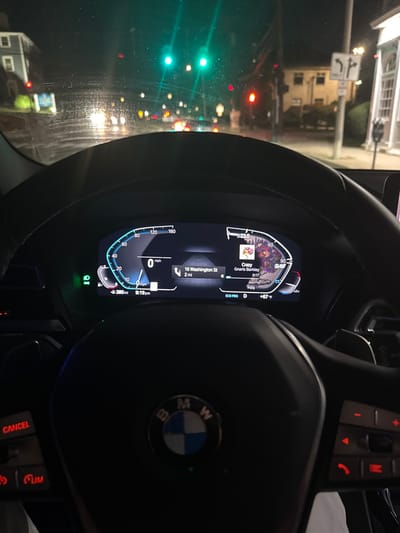 The image provides a driver's perspective from inside a BMW vehicle, stationary at an intersection on Washington Street in Newton, Massachusetts, United States, at 9:19 PM. The digital instrument cluster displays a speed of 0 mph, an external temperature of 67°F, and indicates the vehicle is in "ECO PRO D" mode. Navigation is active, showing the destination "16 Washington St" is 2 miles away. The song "Crazy" by Gnarls Barkley is currently playing. Through the windshield, which has visible smudges or water droplets, multiple traffic lights are illuminated ahead, with two prominent green lights in the center-left and a red light to the right. Distant vehicle headlights are visible. Buildings line both sides of the street, including a light-colored multi-story residence on the left and a building with architectural details on the right. A white traffic sign indicating "ONLY" with left and right turn arrows is visible on the right side of the road. The vehicle's headlights are active, as indicated by a green light on the dashboard. The BMW steering wheel and its integrated controls (cruise control on the left, media/phone on the right) are prominent in the foreground.