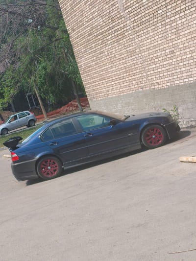 The image depicts a dark blue BMW sedan with red rims parked on an asphalt surface, likely a street or parking lot, in front of a brick building. The car features a noticeable rear spoiler and side skirts. In the background, to the left of the BMW, a silver hatchback is visible, suggesting it might be a parking area or a street with other vehicles. The environment appears to be outdoors during the daytime, with trees and foliage present. The brick building suggests an urban or suburban setting. The overall scene is static, showing parked vehicles and the surrounding environment without any apparent activity. The location is identified as Alchevsk, Ukraine.