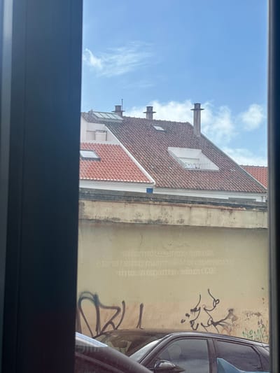 The image displays an urban view observed through a window, framed by dark vertical elements on the left and right. The upper section features a clear blue sky with sparse, wispy white clouds. Below the sky, multiple red-tiled rooftops are visible, indicative of the architecture in Ericeira, Portugal. These roofs are equipped with several chimneys, including cylindrical designs with caps, and numerous skylights. One large, recessed rectangular opening on a prominent roof section appears to contain a faint string of lights or small decorative elements. Below the rooftops, a weathered, light-colored building wall is present, exhibiting faded, mirrored textual reflections and dark graffiti, including an abstract heart-like shape, near its base. The bottom portion of the image shows the dark upper sections of several parked vehicles, partially obscured by the wall and the foreground window frame. No individuals or discernible actions are present in the scene.