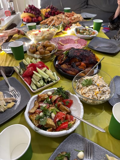 A diverse spread of food is arranged on a yellow tablecloth, likely for a gathering. The dishes include a whole roasted duck, a bowl of sliced tomatoes and cucumbers, roasted potatoes, a salad in a glass bowl, a charcuterie board with various sliced meats, and a pizza in the background. Several green paper cups are placed around the table. In the foreground, a person's hands, adorned with rings, are visible reaching towards the food. The setting appears to be indoors, with natural light illuminating the scene. The presence of a sofa in the background suggests a home environment. The location is identified as Sapronovo, Russia, suggesting this is a domestic meal.