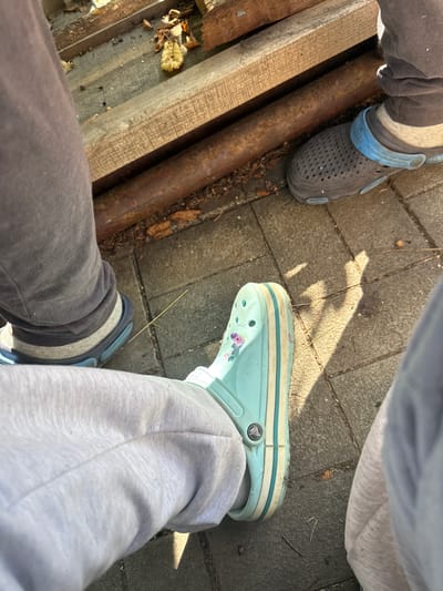 In Balivka, Ukraine, a close-up, high-angle shot captures a ground-level view of several individuals' feet and lower legs, standing on a paved surface. The setting appears to be outdoors, with wooden planks and a metal pipe forming a rustic background. The focal point is on the footwear, with two pairs of Crocs visible. One pair is a dark grey with a bright blue strap, and the other is a light aqua color adorned with small floral patterns. The individuals are dressed in casual attire, including grey sweatpants and socks. The lighting suggests a sunny day, casting distinct shadows on the pavement. The overall scene conveys a relaxed, informal atmosphere.