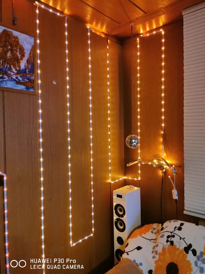 A corner of a room, likely a living room or bedroom, is illuminated by strings of small, warm-toned LED lights that outline the ceiling and descend along the walls. The walls are paneled with wood that has a warm, orange-brown hue, and the ceiling appears to be made of similar material.

On the left wall, a framed painting depicting a landscape with trees and water hangs. Below it, and extending down from the ceiling, is a vertical string of the same LED lights.

In the corner itself, a small, reflective disco ball hangs from a string, catching the light. A thin branch-like object with some lights attached to it is mounted on the wall below the disco ball. To the right of this, a tall, white speaker stands on the floor.

In the foreground, a portion of a couch or chair is visible, covered with a white fabric featuring a floral pattern in shades of orange and black. The overall impression is one of cozy, ambient lighting.

There is no indication of the location being in Bad Oeynhausen, Germany.