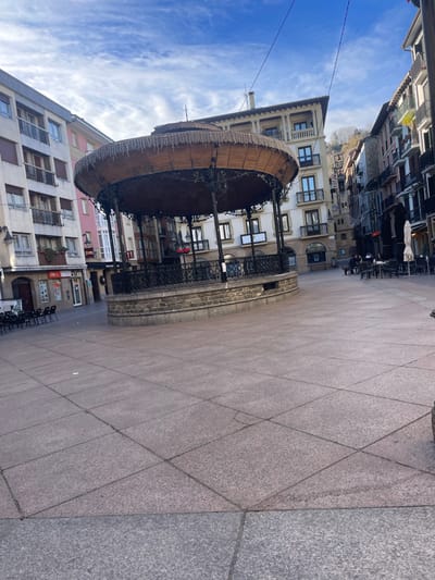 The image depicts the Musika Plaza (Music Square) in Zarautz, Spain, on an overcast day with patches of blue sky visible. In the center of the square stands a circular, elevated bandstand with a dark metal railing and support columns, topped by a light brown, thatched or woven wooden roof structure adorned with string lights or decorative fringe. The bandstand rests on a stone base.

The plaza floor is paved with large, rectangular, light-colored granite-like tiles. To the left of the bandstand, an area with numerous empty black chairs is visible, suggesting an outdoor seating area. To the right, another outdoor seating area is present, with several black tables and chairs, occupied by approximately four individuals. One white umbrella is visible above these tables.

Surrounding the plaza are multi-story buildings. On the left, a light-colored building with multiple balconies and dark window frames rises, showing commercial establishments at ground level. Further into the background, other buildings of varying colors, including pink and white, are visible. On the right, older-style buildings with balconies and dark facades line a narrow street leading away from the square. A distant hillside covered in greenery is visible behind the buildings on the right. Overhead, dark utility wires crisscross the sky. No direct interactions between people are clearly discernible. The scene suggests a quiet, daytime setting in a town square.