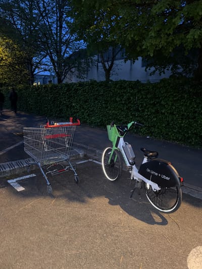 The media file captures an outdoor scene at dusk or night, characterized by low light and artificial illumination. The image is rotated 90 degrees counter-clockwise, presenting the ground plane vertically on the right side and the sky on the left.
The primary subjects are an electric bicycle and a metal shopping cart, both situated on an asphalt surface with faint white painted markings. The e-bike, featuring a prominent lime green and white frame, is equipped with reflective strips on its wheels, which glow brightly in the dim light. Text on the bike's rear fender clearly reads "On Lime + Uber," and "Lime" is visible on its frame, identifying it as part of a shared mobility service. A battery pack is discernible on its frame.
Adjacent to the e-bike, a standard shopping cart with a red handle and red plastic trim is visible. Shadows cast on the asphalt suggest an artificial light source, likely a street lamp, is illuminating the area from an angle.
In the background, a dense wall of dark green foliage, possibly bushes or ivy-covered fencing, extends along what appears to be a property line. Further in the background, to the left (or upwards in the actual orientation of the scene), dark tree silhouettes are visible against a deep blue night sky, with parts of a building faintly seen through the branches. The overall atmosphere is quiet and still, typical of a suburban or semi-urban street at night in Bures-sur-Yvette, France.