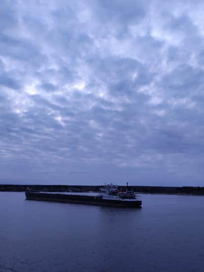 The image shows a cargo ship on a body of water, likely a river or large lake, under a cloudy sky. The ship, labeled "BOAT O-DON 5017", appears to be in transit. The background includes a dark treeline suggesting a shoreline or forest area. The location is likely near or within the city of Oktyabr'skiy, Russia based on the ship's name. The water's surface is relatively calm with minimal ripples. The overall scene is dimly lit, suggesting it is dusk or dawn.