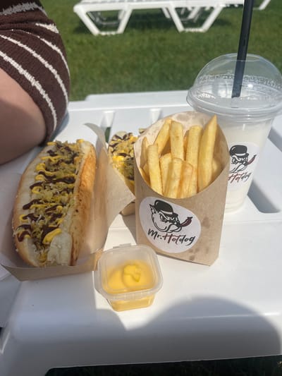 In the city of Emelyanovskoye Lake, Russia, an outdoor setting shows a white table laden with food and drink. On the left, a hot dog sits in a brown paper tray, topped with yellow mustard, a dark sauce, and crispy toppings. Next to it, a clear plastic container holds a yellow dipping sauce. In the center, a brown paper cone is filled with golden french fries. Behind the fries, a clear plastic cup with a domed lid and a black straw contains a white beverage, possibly a milkshake or smoothie. A logo sticker featuring a cartoon pig wearing a hat and smoking a pipe, with the text "Mr. Hotdog," is visible on the fries cone and the cup. In the background, green grass and a glimpse of white lounge chairs suggest a recreational area. A person's leg, wearing brown and white striped shorts, is partially visible on the far left. The scene is bathed in bright sunlight, casting shadows on the table.
