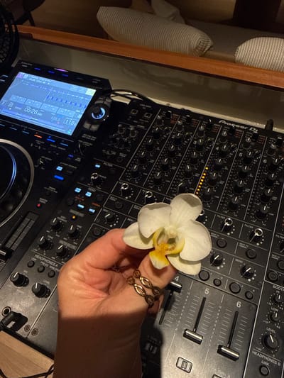 This media file shows a close-up, high-angle shot of a DJ mixer and a white orchid. The mixer, identified by the "Pioneer DJ" branding, has numerous knobs, faders, and buttons. A screen on the mixer displays track information, including "Jazzonita (Original Mix)," a timestamp "05:26," BPM "123.0," and "05:26.599." The scene appears to be indoors, with wooden elements visible in the background, suggesting a studio or entertainment space. A hand, adorned with two rings, is holding the orchid, which is positioned over the mixer. The lighting suggests it is nighttime or a dimly lit environment. The overall mood is calm and artistic, with the juxtaposition of the technical DJ equipment and the delicate flower.