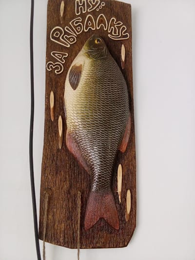 A carved wooden plaque depicting a fish is mounted on a white wall. The plaque is rectangular with rounded corners and features a textured, dark brown wood surface. A three-dimensional carving of a silver-scaled fish with reddish fins and tail is centered on the plaque. Above the fish, carved in stylized Cyrillic letters, are the words "НУ, РЫБАЛКЕ!" which translates to "Well, fishing!". The text is also carved and painted in a light brown color. On either side of the fish, near the top of the plaque, are small, carved oval shapes resembling wooden pegs. Two lengths of rough, tan twine hang from the bottom of the plaque, suggesting it is intended for hanging. To the left of the plaque, a thick black cable is visible, running vertically along the wall. The overall scene appears to be an interior setting, possibly a home or a decorative display. No people are present in the image. The location in Altai, Kazakhstan, is not specified by any visual cues in the image.