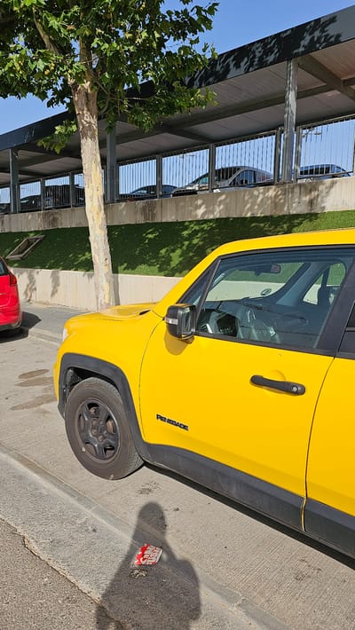 The image displays a bright yellow Jeep Renegade parked on a paved surface, likely a street or parking area, with a curb separating it from a grassy area and a covered walkway. The walkway is supported by concrete pillars and has a metal railing, behind which other vehicles are partially visible. A mature tree with light-colored bark and green leaves stands to the left of the Jeep, casting dappled shadows on the ground. The sky is clear and blue, suggesting a sunny day. A crumpled red and white paper item, possibly a discarded wrapper, is on the ground near the rear wheel of the Jeep. The text "RENEGADE" is visible on the side of the car. The scene appears to be in Cuarte de Huerva, Spain. The orientation of the photo is rotated.