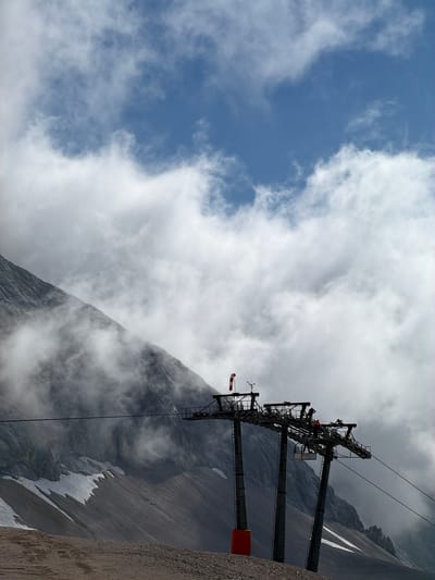 In Wildermieming, Austria, a ski lift station is visible against a backdrop of a cloudy sky and a steep mountain slope. The ski lift structure consists of two large, dark-colored pylons supporting a complex network of wheels and cables. A red and white windsock is mounted on the left pylon, and a few individuals, wearing orange vests, appear to be working on the platform of the ski lift. Below the ski lift, the mountain's rocky terrain is visible, with patches of snow scattered across its surface. The foreground shows a gravelly, uneven ground. The overall scene suggests an operational, yet potentially challenging, environment due to the weather conditions.