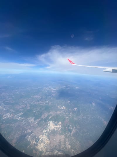 The image, taken from an airplane window, shows the wing of the plane with a red Turkish flag on its tip. Below, the Earth is visible, with a mix of urban and rural landscapes. The sky ranges from clear blue to cloudy white. The image is rotated such that the horizon appears to be vertical. Based on the urban layout and general landscape characteristics visible from the plane window, it is likely that the location in the city of San Vittorino, Italy. is above the landscape.