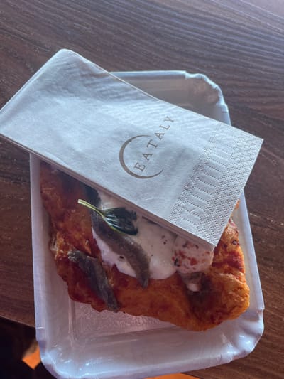 An overhead shot captures a single serving of an Italian dish, likely a fried pizza (pizza fritta), placed on a white, rectangular, disposable paper tray with slightly raised edges. The dish features a golden-brown, puffy, fried dough base, topped with white dollops of what appears to be mozzarella or burrata cheese, a few dark green basil leaves, and several silvery anchovy fillets.
A folded white paper napkin rests partially over the food on the left side of the tray. The napkin has a textured, patterned border along its top edge and prominently displays the word "EATALY" in a brown, sans-serif font, enclosed within a minimalist circular design.
The tray and its contents are situated on a dark, wood-grained surface, possibly a table or countertop. The lighting is even, suggesting an indoor setting, likely during the daytime. No people are visible, and the scene depicts a prepared meal ready for consumption in a casual dining environment, possibly within an Eataly food hall or similar establishment. The focus is entirely on the food and its presentation.