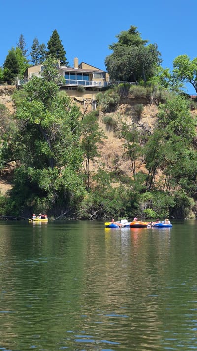 A group of people are seen relaxing on inflatable tubes in a body of water. The scene is set against a backdrop of lush green trees and a hill with a house on top. The water appears calm with reflections of the surrounding trees. The house is a light tan color with large windows and a balcony. The sky is a clear blue. There are several individuals scattered across the water, each in their own inflatable tube. Some appear to be lying down, while others are more reclined. The individuals are dressed in casual summer attire. The overall impression is one of leisure and outdoor recreation. This scene is likely in Carmichael, California, given the style of housing and vegetation, characteristic of the area.