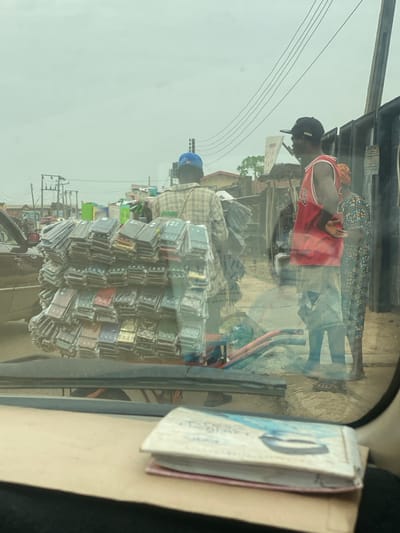 The scene depicts a street in AlakIa, Nigeria, viewed from inside a vehicle under an overcast sky. In the foreground, a light-colored vehicle dashboard with a small booklet is visible. The midground features a male vendor, observed from behind, wearing a blue baseball cap and a plaid shirt, positioned behind a blue and red metal pushcart. The cart is heavily loaded with numerous stacks of clear plastic-wrapped mobile phone cases, displaying various colors and designs. To the right, a second male stands in profile, wearing a black baseball cap, a red sleeveless jersey with the number 23, and light-colored shorts; his right hand rests on his lower back. Partially obscured behind him is a female in a patterned garment. The background reveals power lines, utility poles, a distant traffic signal, and rudimentary structures lining an unpaved road or dirt path.