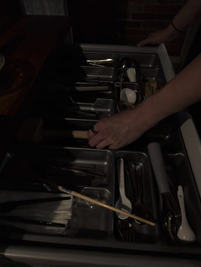 The image shows a close-up of a person's hand reaching into a kitchen utensil drawer. The drawer is a standard organizer with compartments for different types of cutlery. Several utensils are visible, including spoons, forks, and knives, as well as some white-handled items. The hand, adorned with a ring on the ring finger, is gripping a wooden-handled mallet, possibly a meat tenderizer. The drawer itself appears to be made of gray plastic, and the background suggests a kitchen setting with brickwork visible on the left. The lighting is dim, creating shadows and highlighting the metallic sheen of the cutlery. The overall impression is that the person is in the process of selecting or retrieving a utensil from the drawer. There is no visible text in the image. The context of Słupsk, Poland is noted but not visually discernible from the image itself.