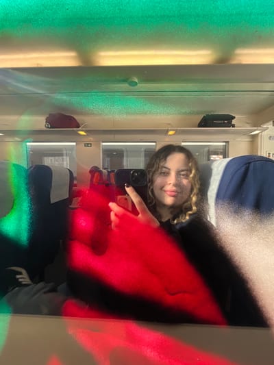 The image depicts a selfie taken within a passenger train carriage, observed through a reflective surface, consistent with a location in the city of Tortosa, Spain. A young female subject with curly, light brown hair and blonde highlights, wearing a dark top, is centered in the reflection. She holds a dark-colored smartphone with a multi-lens camera system in her right hand, capturing the image. Her left hand is also visible, displaying dark nail polish. She exhibits a slight smile. The train interior features rows of dark-upholstered seating with white headrest covers. Overhead luggage racks contain a reddish-brown backpack on the left and a dark bag on the right. Fluorescent strip lighting illuminates the ceiling, which also features a white circular fixture. Intense red and green light reflections, originating from an unseen external source or internal interaction with the reflective surface, dominate the foreground and midground, casting strong color across the scene and partially obscuring the lower half of the subject and parts of the train interior. Feet wearing light socks and dark shoes are visible in the lower-left foreground. Further sections of the train carriage are visible through interior glass partitions in the background.