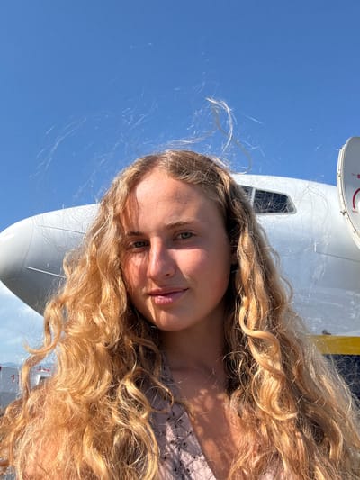 This is a portrait of a young woman with long, wavy blonde hair, taken outdoors. She is looking towards the camera, with her face partially obscured by her hair. Her eyes are green, and she has a slight smile.
Behind her, a white aircraft, possibly a small plane or helicopter, is visible. Part of the aircraft's fuselage and a window are in the frame. The sky is a clear, bright blue, suggesting a sunny day. A yellow stripe is visible on the aircraft.
The image appears to be a selfie, with the camera held at an angle to capture both the woman and the aircraft. The lighting suggests it's daytime. The overall mood is casual and relaxed. No visible text is present in the image. The context indicates the location is Kruja, Albania.