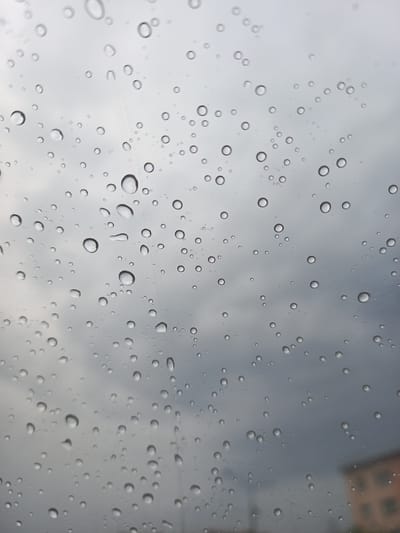 This is a close-up, angled shot of raindrops on a windowpane, with a muted, cloudy sky visible through the glass. The raindrops vary in size, from tiny droplets to larger, more defined spheres. Some are perfectly round, while others are slightly elongated or irregular, suggesting movement or merging. The overall impression is one of a cool, overcast day, possibly during or after a rain shower. The light is diffuse and even, characteristic of cloudy weather. There are no discernible people or specific objects within the frame, apart from the water droplets themselves, which are the primary subjects. The image evokes a sense of calm, perhaps even melancholy, due to the subdued color palette and the common association of rain with introspection. The background sky is a mix of light and darker grays, with subtle cloud formations visible, indicating a natural, outdoor setting. The time of day is ambiguous due to the lack of direct sunlight. The scene is generic enough to be almost anywhere experiencing rain, but the provided location context of Norilsk, Russia, imbues it with a sense of Northern latitude and potentially colder climes. There is no visible text in the image.