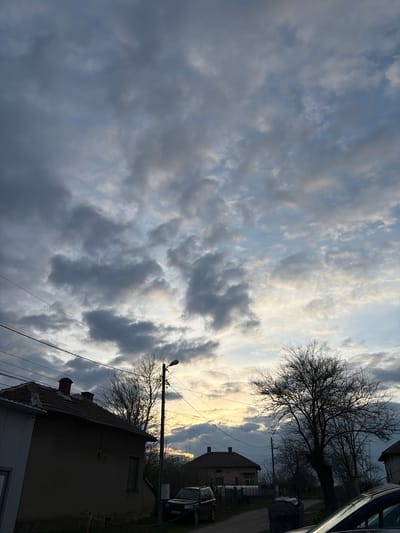 The image captures a low-angle view of a cloudy sky in Valchedram, Bulgaria. The sky is filled with cumulus clouds, ranging in color from dark grey to pale white. A bare tree, likely in its dormant stage, stretches across the upper right portion of the frame, silhouetted against the sky. In the lower right, the corner of a building is visible, featuring a tiled roof and a small chimney. Power lines run across the frame, intersecting with the tree branches. In the background, a portion of another building can be seen, partially obscured by the tree and other objects. A car is parked beside the upper building. The lighting suggests either early morning or late afternoon.