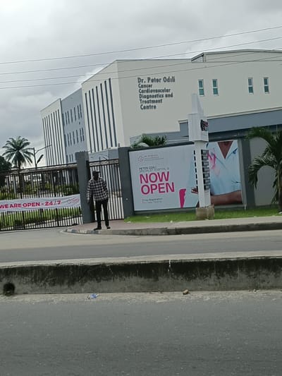 The image shows the exterior of the Dr. Peter Odili Cancer Cardiovascular Diagnostics and Treatment Centre in Rumuepirikom, Nigeria. A man in a checkered shirt and dark pants stands near a gated entrance. A banner on the gate reads "WE ARE OPEN - 24/7". A large advertisement on a billboard to the right displays "PETER ODILI SPECIALIST HOSPITAL IS NOW OPEN" with "Free Registration For New Patients" at the bottom. The building is modern with a light-colored facade and vertical grey accents. Palm trees and other foliage are visible in the background. A road with a concrete barrier runs in the foreground. The sky is overcast.