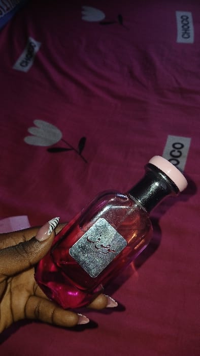 A dark-complexioned hand holds a perfume bottle, angled against a magenta textile background. The hand displays manicured nails; two visible nails feature a white base with black zebra stripes, while others have a nude base with white tips. The transparent glass perfume bottle contains a dark pink liquid and bears a silver metallic label inscribed with black Arabic script, appearing to read "وصوف". The bottle's neck is black, capped by a light pink top. The background textile, consistent with a bedsheet, is magenta and patterned with repeating white tulip-like flowers and vertically oriented white rectangular tags displaying the capitalized text 'CHOCO'. The fabric is wrinkled, and the scene is under dim illumination. No visual cues within the image confirm its specific location in the city of Igbede, Nigeria.
