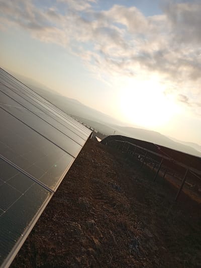 The image displays a solar farm located in Drugan, Bulgaria, captured during either sunrise or sunset. In the foreground and extending to the mid-ground on the left, a long array of dark, rectangular photovoltaic solar panels is prominently featured, angled upwards. Reflections are visible on the panel surfaces, with slight irregularities and possible moisture droplets along their lower edges. The immediate ground area underneath and between the panel rows consists of dry, disturbed brown earth, indicating recent activity or sparse, low vegetation. Further down the slope in the mid-ground on the right, additional solar panel structures are visible in partial shadow, identifiable by their dark frames and support posts. The background presents a series of hazy, layered hills or mountains extending towards the horizon. The sky, which occupies the upper half of the image, is light blue with scattered white and grey clouds, particularly dense on the right. A strong, bright sun is positioned low on the right side of the horizon, emitting a significant glare and illuminating the scene with warm, golden light. No individuals, active interactions, or specific equipment beyond the solar installation are present in the frame.