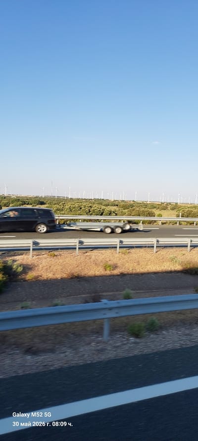 The image shows a highway with a black van driving on it. Adjacent to the highway is a field populated with wind turbines. The date and time stamp on the image is "30 Maí 2026 г. 08:09 ч." The location is likely in the vicinity of Tébar, Spain, based on the presence of wind turbines in the landscape, as wind turbine farms are common in that region of Spain.