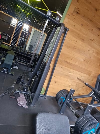 The image depicts the interior of a fitness facility located in Oberseelbach, Germany. The scene is static, devoid of individuals.

In the foreground, a dark grey padded seat of a gym machine is visible. To its right, a dark grey leg press machine is equipped with four circular, dark grey weight plates, each featuring blue accents and likely indicating a 25kg mass, stacked evenly on both sides. The floor is covered with dark grey rubber mats.

In the midground, a prominent dark grey cable machine stands, featuring visible weight stacks and multiple pulleys and cables. A black double-ended rope attachment and a folded grey towel are situated on the rubber mat floor near the base of this machine.

The background to the left features a large mirror reflecting another section of the gym. Within the reflection, tiered racks holding numerous dumbbells of varying weights are visible, along with a black padded bench. A window with a light frame, partially obscured, is also reflected. The wall behind the reflected equipment appears dark, possibly textured brick.

The wall on the right side of the image is clad with light brown wooden panels. The ceiling is dark, fitted with distinct linear yellow light fixtures. A segment of a light green wall or structural beam is visible at the junction of the wooden wall and the ceiling.