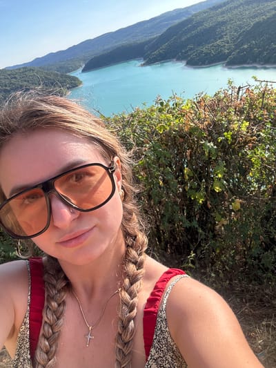 A young woman is captured in a selfie against a backdrop of a scenic turquoise lake surrounded by lush green mountains under a clear blue sky. She is wearing large, orange-tinted sunglasses, a patterned dress with red ruffled straps, and a gold necklace with a cross pendant. Her blonde hair is styled in two braids. The image appears to have been taken outdoors, likely in a natural setting with dense vegetation in the foreground. The location is identified as Moirans-en-Montagne, France.