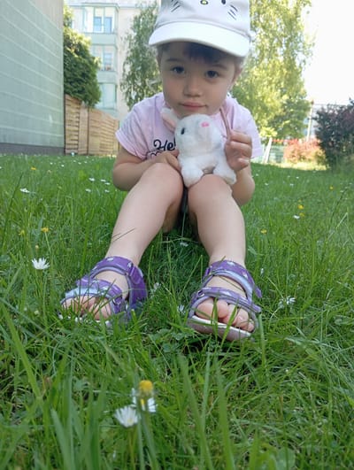 In Radzymin, Poland, a young girl wearing a white cap with a cartoon cat face and a pink t-shirt sits on a grassy lawn. She is holding a small, white stuffed rabbit with pink accents. The girl is wearing purple sandals with a geometric pattern. The background shows a building, a wooden fence, and trees. The focus is on the girl and her toy, with the grass and flowers in the foreground.