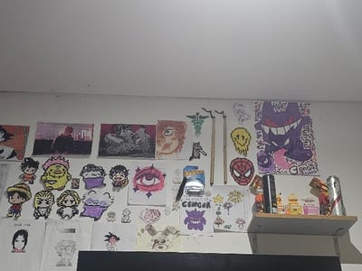 A wall is covered with an assortment of drawings, prints, and pixel art. Anime characters such as Goku and Luffy are visible. There are also drawings of Pokemon, notably Gengar, and a pixel art Spider-Man. A small shelf holds various items including cans, small toys, and figurines. The overall impression is a collection of artwork and memorabilia.