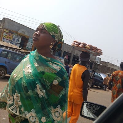 The image depicts a vibrant street scene in Olomoda, Nigeria, under a bright, clear sky. In the left foreground, a woman with dark skin is shown from the chest up, looking slightly upward and to the right. She wears a green patterned headwrap and a green patterned garment, layered with a sheer green fabric adorned with white floral embroidery. Gold-colored hoop earrings are visible on her ears. To her left, the front portion of a blue car is parked. In the midground, a man with dark skin walks away from the camera, balancing a large, round silver tray piled with numerous light brown baked goods on his head. He is dressed in an orange tank top and matching orange trousers, with a dark band around his head. Other individuals are partially visible further down the street, including one on the far right wearing an orange and brown patterned garment. The background features a street lined with single to two-story buildings, some with corrugated metal roofs and visible signage, including "OLAS" and a partial "Nimi" sign. Overhead electrical wires are strung across the scene. Vehicles are present on the street, suggesting an active commercial area.